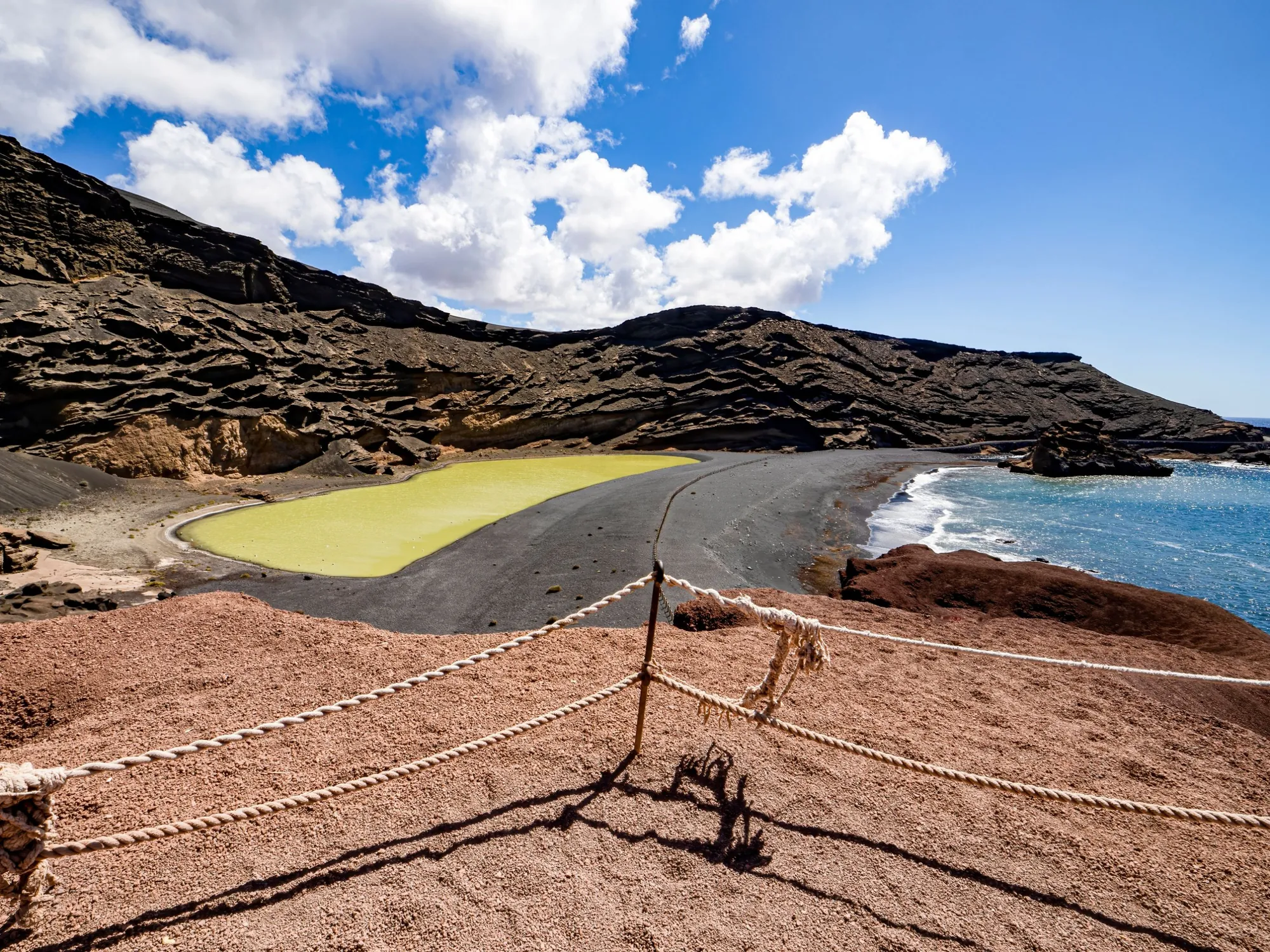 A rope fence above a green lagoon and a black sand beach, with layered dark cliffs and the sea